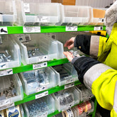 A green metal trolley racking unit with multiple shelves, holding white plastic bins